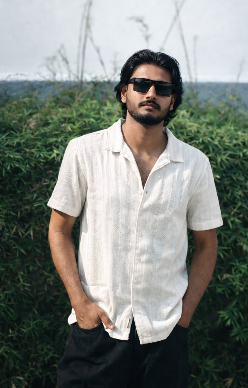 Portrait of Ajas Muhammed in a white shirt and sunglasses, standing in front of a green hedge